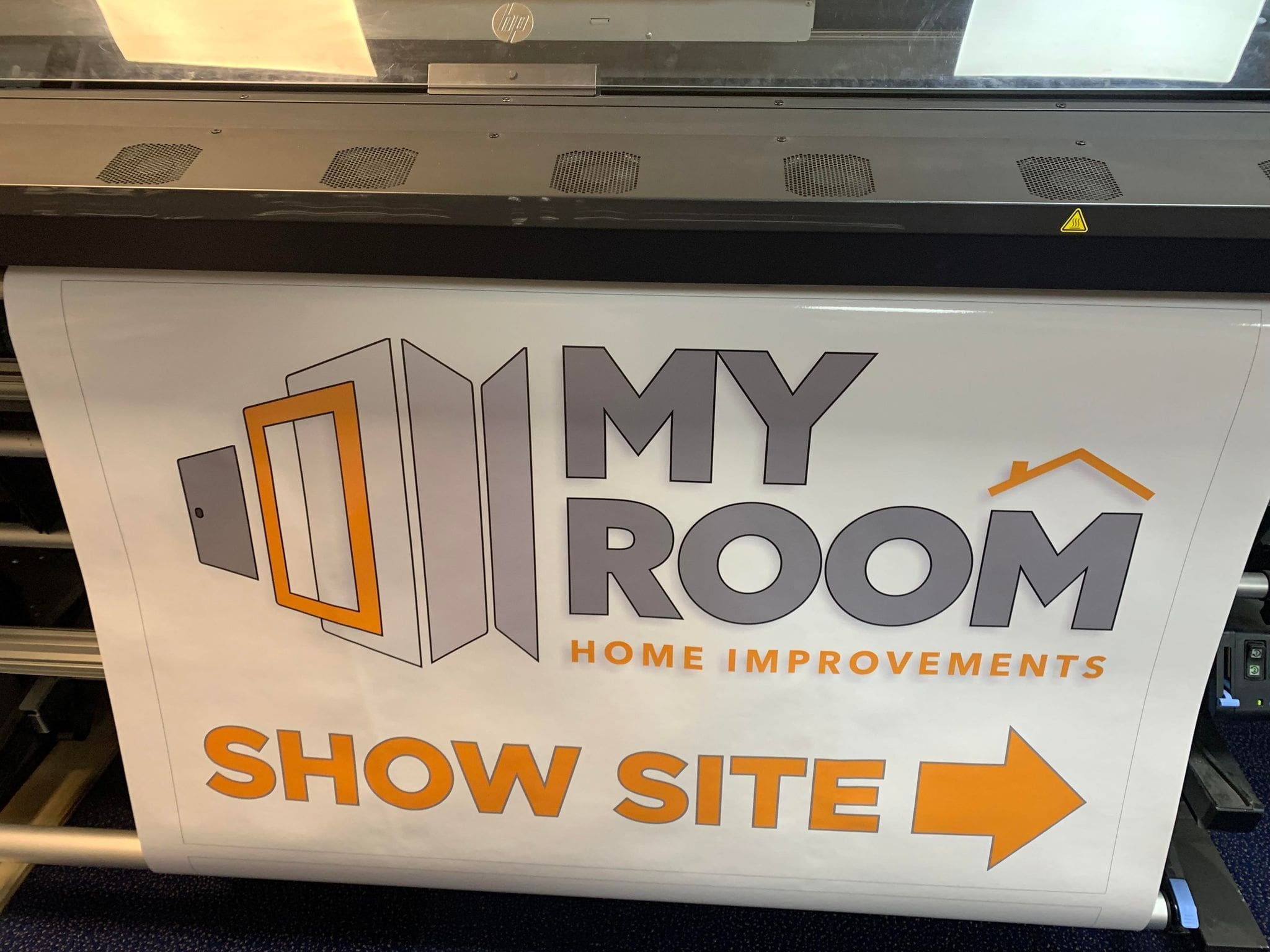 A large format printer is printing a banner.   The banner is white with orange and gray graphic elements.  The words 