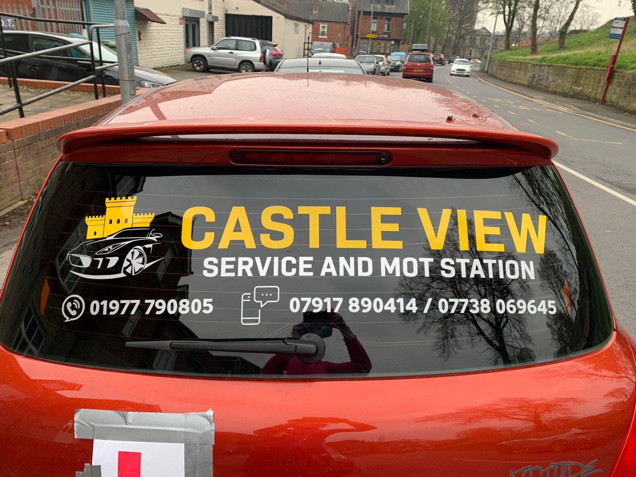 Full rear view of an orange/red car with a large, black vinyl sticker on the rear window. The sticker displays the words 