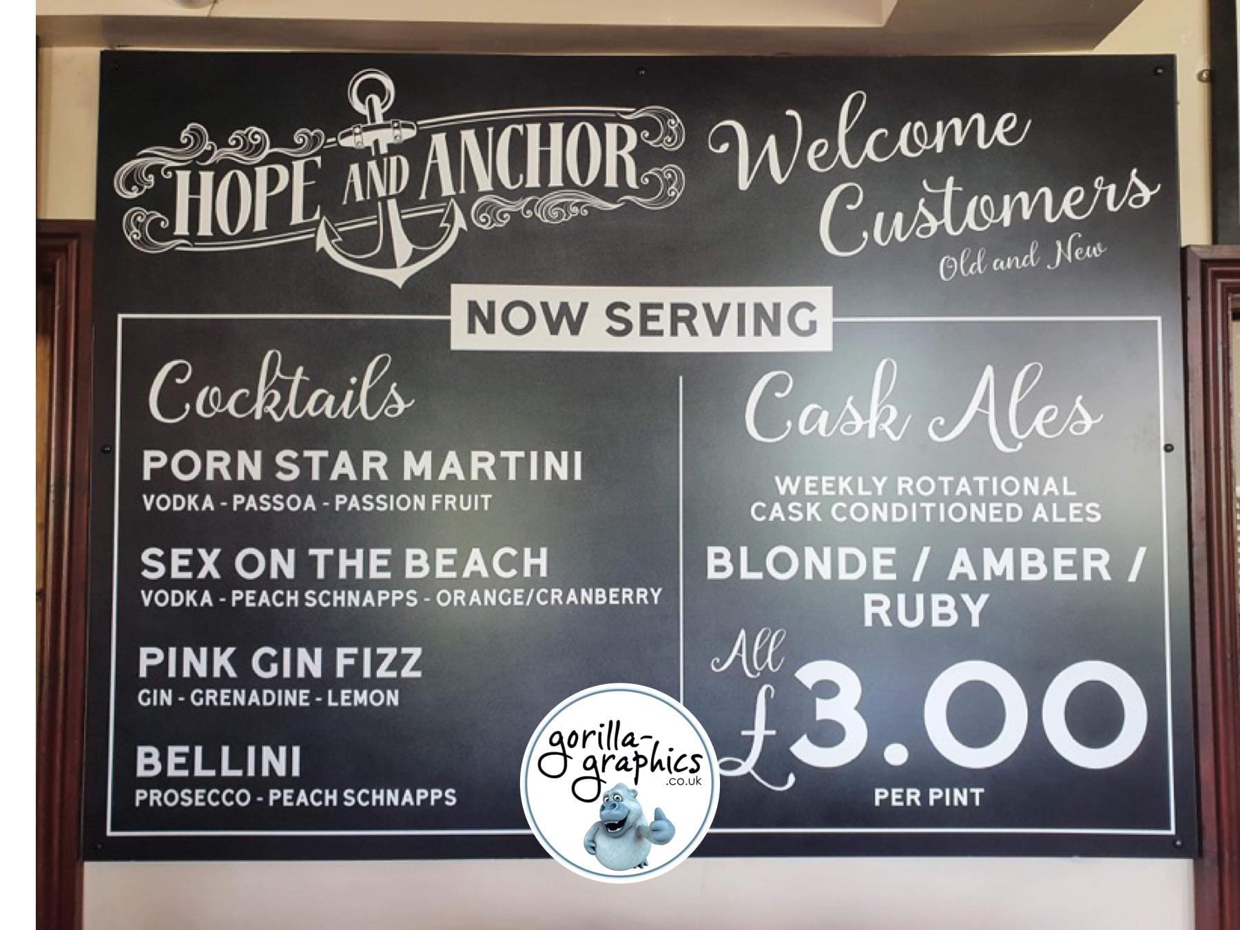 Black chalkboard-style sign with white lettering advertising cocktails and cask ales.   The top of the sign features the words 